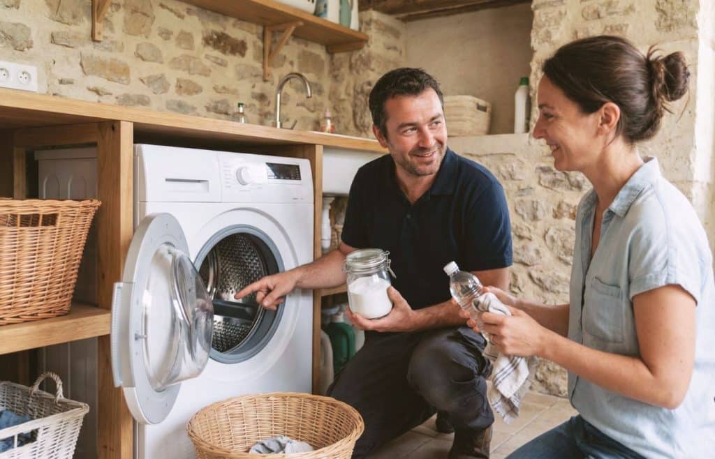 Un réparateur professionnel (Damien) montre à Elise Durant comment utiliser du bicarbonate de soude et du vinaigre blanc pour entretenir et détartrer une machine à laver dans une buanderie au style rustique.